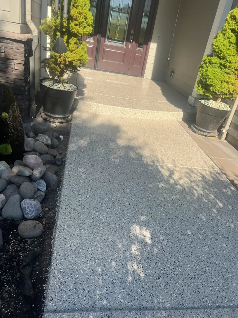 An Oregon-inspired concrete pathway with a speckled texture leads to a welcoming entrance with double doors, flanked by potted plants and small rocks. The durable concrete coating ensures longevity and charm in any season.