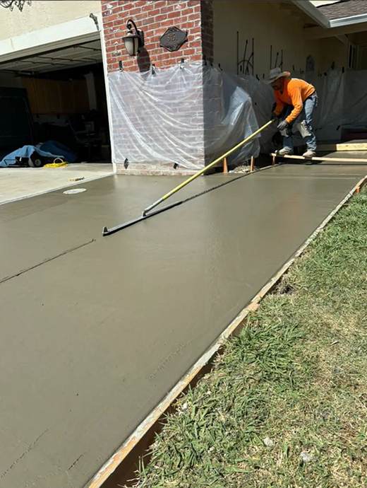 In Oregon, a worker in an orange shirt is expertly smoothing wet concrete on a driveway, using a long tool. Grass edges the scene, while plastic sheeting protects a brick wall. This meticulous process ensures a durable finish perfect for any Concrete Coating application.