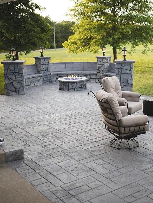 A serene stone patio with a cushioned chair, fire pit, and stone seating offers a cozy retreat. Surrounded by Oregon's lush greenery and enhanced with durable concrete coating, it's the perfect outdoor haven.