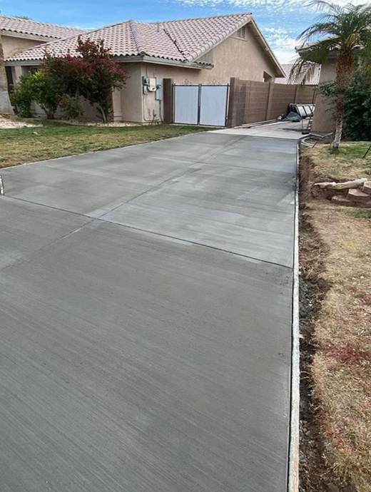 A freshly poured concrete driveway in an Oregon residential area, adjacent to a beige house with a tiled roof, is beautifully bordered by a lawn and small palm tree. Consider adding a concrete coating for enhanced durability and style.