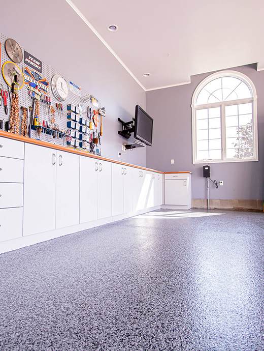 The immaculate garage features a sleek speckled gray concrete coating, complemented by white cabinets and a pegboard wall organizing the tools. Under the large arched window, an Oregon sunset graces the flat-screen TV mounted on one wall.