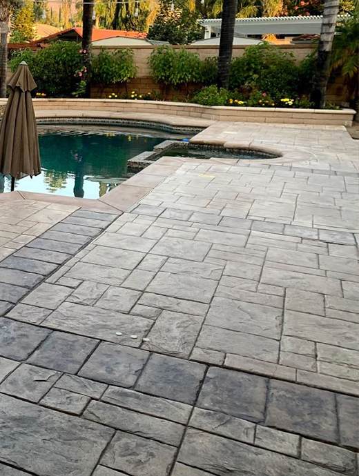Stone-patterned patio with concrete coating leads to a pool with an umbrella stand nearby, surrounded by Oregon's lush greenery and a wooden fence in the background.
